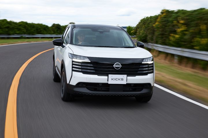 白系ボディのキックスがカーブした舗装道路を左カーブに沿って右向きに走行している。車両は斜め前方から捉えられ、背景には緑の植生と曇り空が広がる。
