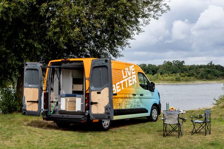 Blick von schräg hinten auf einen grün-orangen Nissan Interstar mit PlugVan Wohnmodul, das man durch geöffnete Hecktüren sieht. Das Fahrzeug steht unter einem Baum auf einer grünen Wiese an einem Fluss steht mit Campingmöbeln daneben