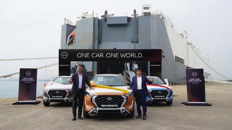Two men in business suits stand in front of a New Nissan Magnite car. In the background is a large container ship which will be transporting the car from India. Two other cars can also be seen in the background.