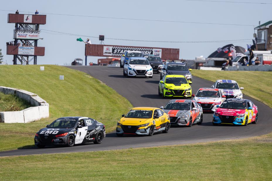 Four race cars are parked side by side on a racetrack with four drivers standing next to them. The cars are colored yellow, gray, and white with various sponsor logos. The drivers are wearing racing suits in colors matching their respective cars.