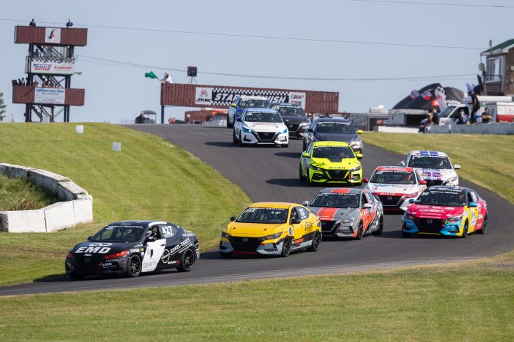 Four race cars are parked side by side on a racetrack with four drivers standing next to them. The cars are colored yellow, gray, and white with various sponsor logos. The drivers are wearing racing suits in colors matching their respective cars.