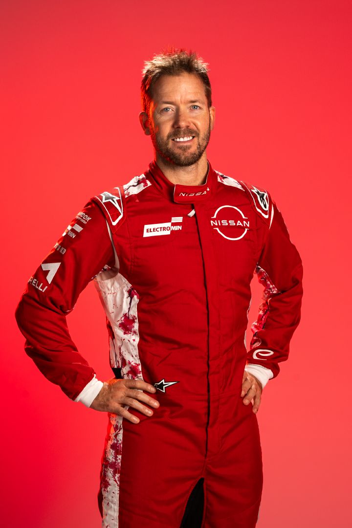 Sam Bird mit den Händen in den Hüften in seinem roten Rennanzug des Nissan Formula E Teams lächelt vor einem roten Hintergrund.