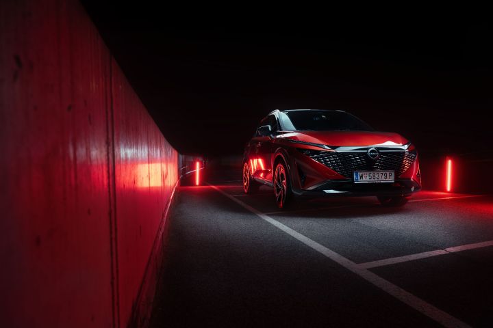 Ein Nissan Qashqai in Fuji Sunset Red steht im Dunkeln auf einem Parkplatz mit roten Leuchtakzenten vor dem schwarzen Hintergrund