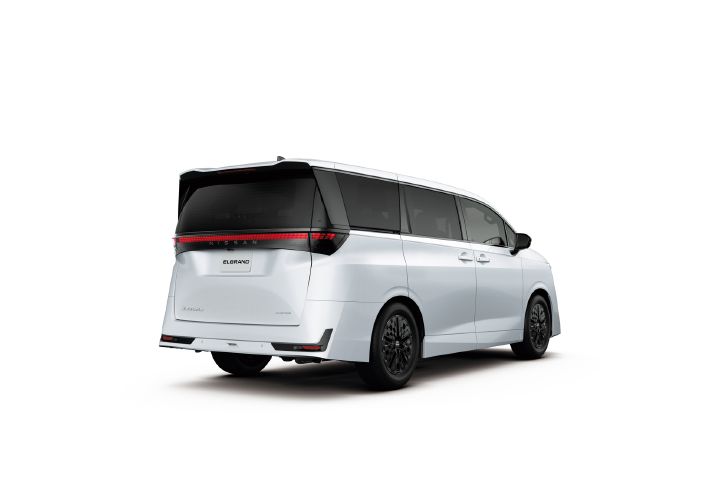 Studio image of the All-new ELGRAND in white, shown from a rear three-quarter view facing right. The vehicle features a black roof and dark window graphics that create a sleek, elongated profile. A full-width LED rear light bar spans the tailgate, with the NISSAN badge positioned at the center. Black alloy wheels and sharply sculpted rear bumper surfaces add a modern, sophisticated appearance. The background is a plain light gray studio setting, allowing the vehicle's design details to stand out clearly.