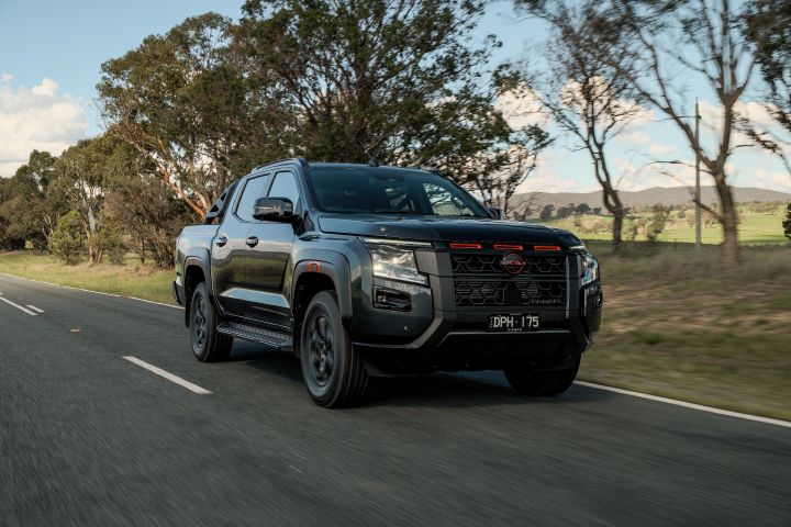 2026 Nissan Navara PRO-4X finished in Boulder Grey portrayed in a dynamic three-quarter road image with blurred roadside trees, dark alloys, sports bar and mountains across the horizon.