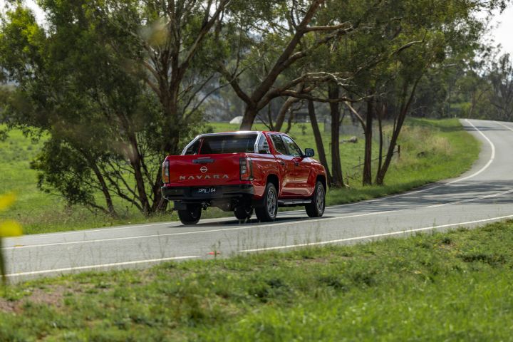 Rear view of a red 2026 Nissan Navara ST‑X driving through a green, tree‑lined country road.