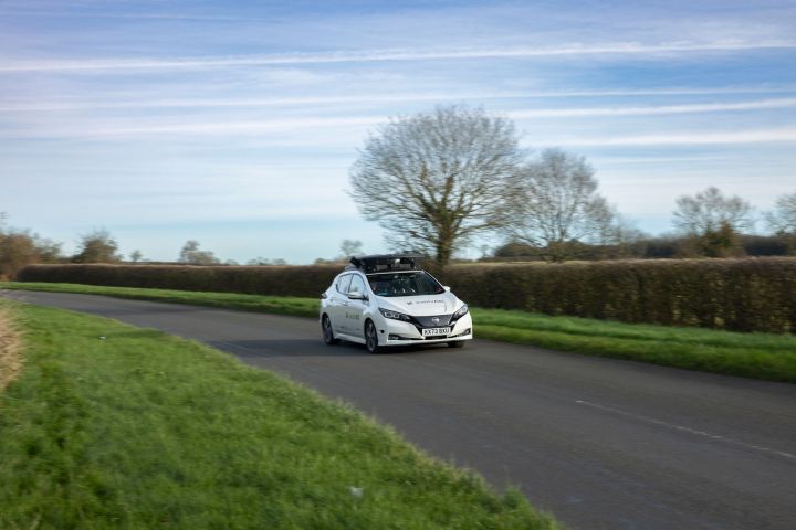 3/4 Frontansicht des evolvAD Nissan LEAF Testfahrzeugs, das auf einer Landstraße zwischen grünen Wiesen und Hecken um eine Kurve fährt