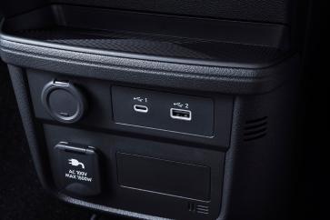 「日産サクラ」のセンターコンソール下部をクローズアップした画像。黒い内装パネルに電源ソケットやスイッチ類が配置され、操作部は正面から見える構図となっている。背景は暗めで、操作部が際立っている。