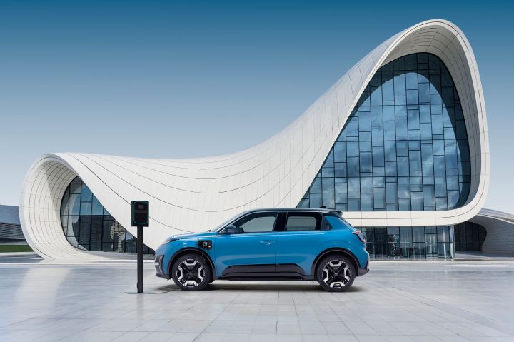 profile view of gear blue body color Micra connected to a charging outlet in front of a design architectural building