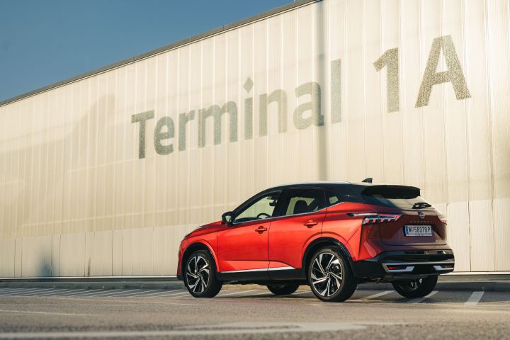 Blick von schräg hinten auf einen Nissan Qashqai in Fuji Sunset Red auf ein Gebäude mit der Aufschrift Terminal 1A