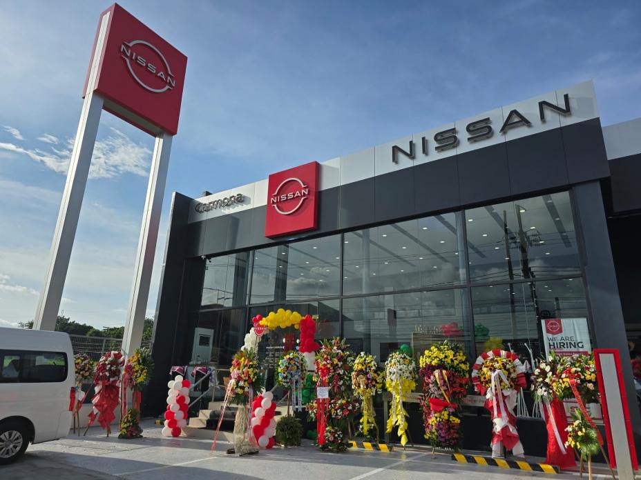 Exterior of the Nissan Carmona dealership with a modern glass facade, featuring a large red and white Nissan logo on the building and a tall signpost. The entrance is adorned with colorful flower arrangements, suggesting a grand opening or special event