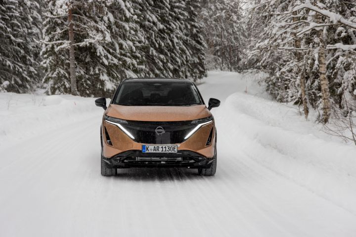 Elektroauto Nissan Ariya e‑4ORCE in Akatsuki Copper mit Dach und Außenspiegel in Pearl Black fährt auf schneebedeckter Straße mit verschneitem Wald auf beiden Seiten, Front