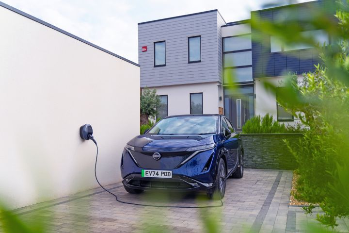 A dark blue Nissan Ariya electric car parked on the driveway of a modern house. The car is connected to a Pod Point charger attached to a white wall.