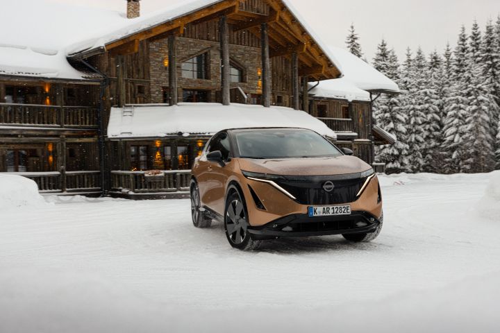 Elektroauto Nissan Ariya e‑4ORCE in Akatsuki Copper mit Dach und Außenspiegel in Pearl Black vor schneebedeckten Haus und Bäumen, 3/4 Front