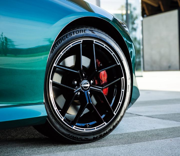 Close-up view of the front-right wheel area of an Unryu Green Fairlady Z. The image focuses on the front-right fender and wheel, highlighting the rich Unryu Green bodywork and its reflective surface. A black multi-spoke alloy wheel dominates the frame, with a red brake caliper visible behind it. The tire sidewall displays the “BRIDGESTONE” branding, while a modern glass-fronted building appears softly blurred in the background. The composition emphasizes the sporty chassis components and premium detailing of the Fairlady Z.