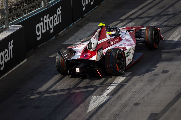 Ein rot-weißer Formel-E-Rennwagen fährt auf einem Straßenkurs, der von Zäunen und Sicherheitsbarrieren gesäumt ist. Im Cockpit ist Oliver Rowland zu sehen, während ein weißer Richtungspfeil und Schatten der Zäune auf die Fahrbahn fallen.