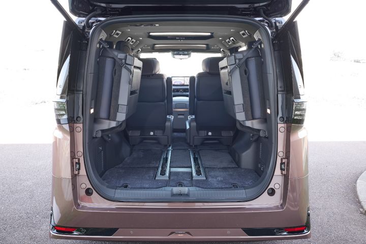 新型エルグランドのリアハッチを開けた状態で、車両後方から車内を正面に捉えたビュー。3列目シートは左右に跳ね上げて収納されており、側面に固定された状態でラゲッジスペースが大きく拡張されている。中央には床面のカーペットと前後に伸びるシートレールが見え、フラットで広い荷室空間が確保されている。前方には2列目キャプテンシートとセンターコンソール、そのさらに奥にダッシュボードと大型ディスプレイが見える。天井には照明やエアコン吹き出し口が配置され、外からの強い日差しで車内全体が明るく照らされている。