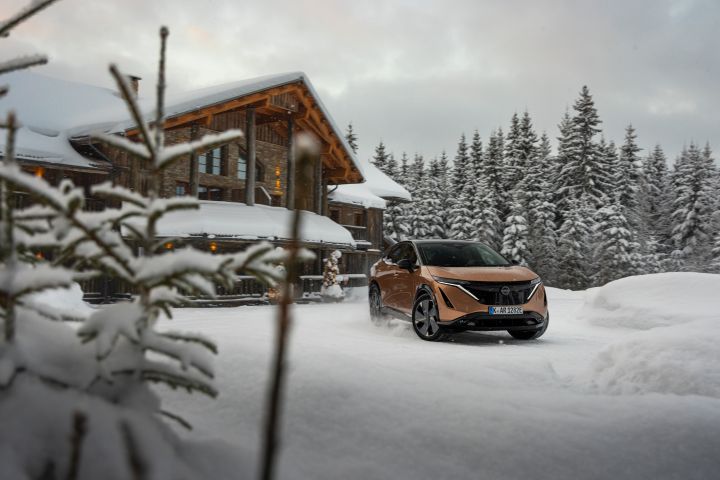 Elektroauto Nissan Ariya e‑4ORCE in Akatsuki Copper mit Dach und Außenspiegel in Pearl Black fährt vor schneebedeckten Haus und Bäumen, 3/4 Front