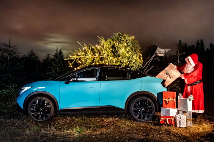 Seitenansicht eines türkisblauen Nissan LEAF, durch dessen geöffnete Heckklappe ein Weihnachtsmann den Kofferraum mit verpackten Weihnachtsgeschenken belädt, auf dem Dach liegt eine Tanne, die mit einer Lichterkette geschmückt wurde