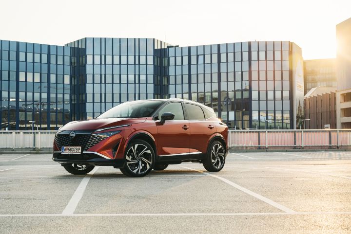 Blick von schräg vorne auf einen Nissan Qashqai in Fuji Sunset Red auf einem Parkplatz vor einem Hotel