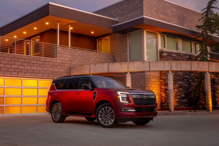 Front right 3/4 of a red 2025 Nissan Armada parked in the driveway of a modern stone mansion at sunset.
