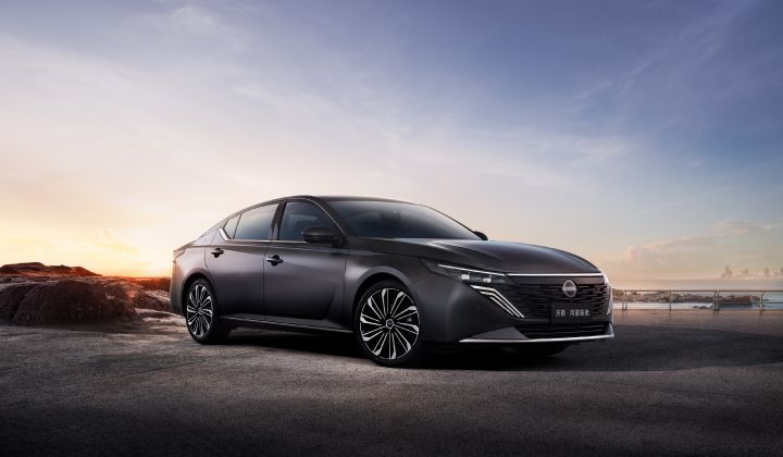 A dark gray new Teana sedan parked on a road with rocky terrain and a coastline in the background. The car faces left, captured from a front-side angle highlighting its wide grille and slim headlights. The backdrop shows a sunset sky transitioning from blue to orange, with distant ocean and rocks visible.