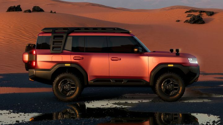 In a desert landscape, a red-orange Terrano PHEV Concept is parked with its right side facing the viewer, illuminated by the light of sunset. Sand dunes and distant mountains extend across the background, while reflections of the scenery and the vehicle appear in a puddle in the foreground.