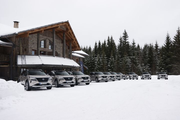 Frontaler Blick auf zehn silberner Nissan X‑Trail e‑Power e‑4ORCE, die im Schnee vor einem großen Holzhaus stehen