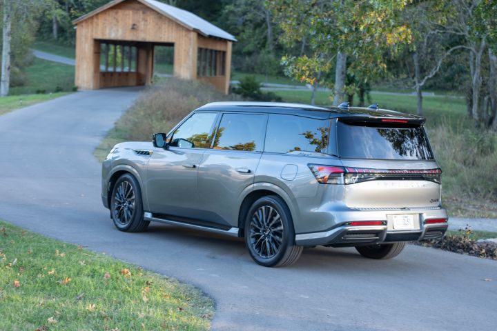 Left rear 3/4 angle of a 2025 INFINITI QX80 parked on an asphalt path by a covered bridge.