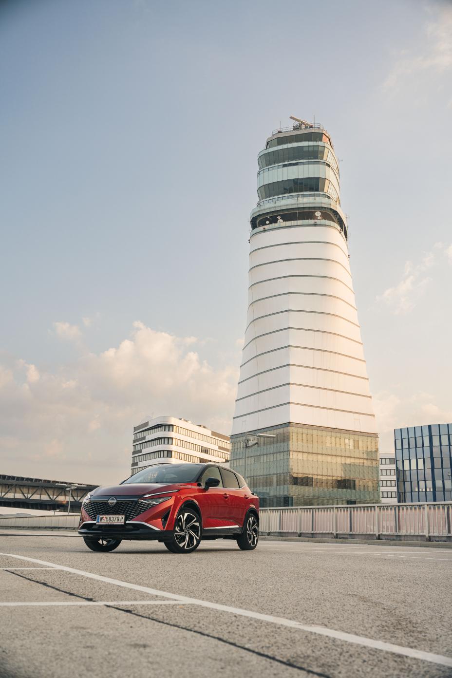 3/4 Frontansicht eines Nissan Qashqai in Fuji Sunset Red auf einem Parkplatz vor einem hohen weißen Turm