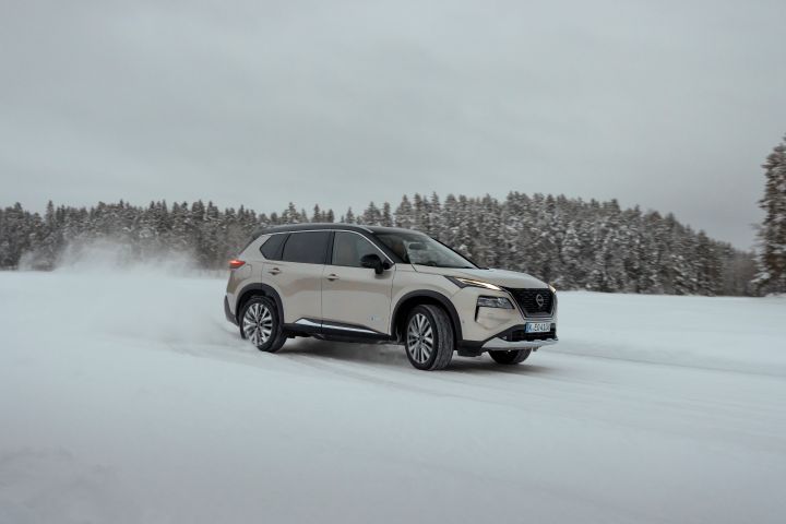 Silberner Nissan X‑Trail e‑Power e‑4ORcE fährt auf schneebedeckter Straße mit verschneiten Bäumen im Hintergrund, 3/4 Front