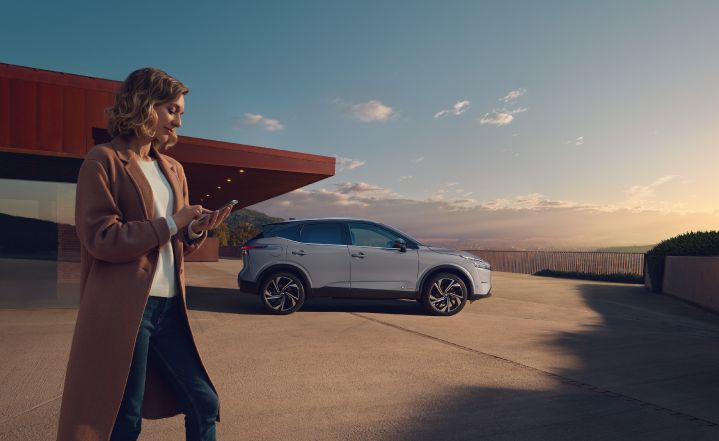 Eine Frau steht vor einem Nissan Qashqai e‑Power in Ceramic Grey und schaut auf ihr Smartphone.