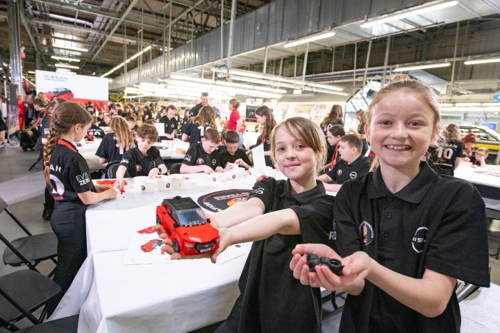 Two school children holding a LEGO Nissan Qashqai, with more school children visible in the background.