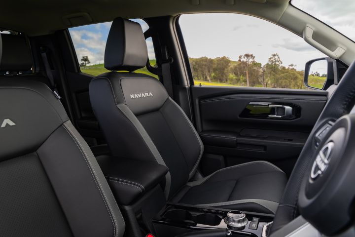 Wide interior seating view of a 2026 Nissan Navara ST-X, showing the front passenger seat, centre armrest, centre console, and part of the driver’s seat, with countryside visible through the side windows.