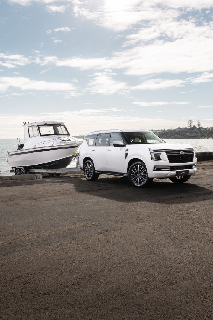 2026 Nissan Y63 Patrol finished in white, shown from a front three-quarter exterior angle on a coastal boat ramp towing a large white boat, with the ocean in the background.