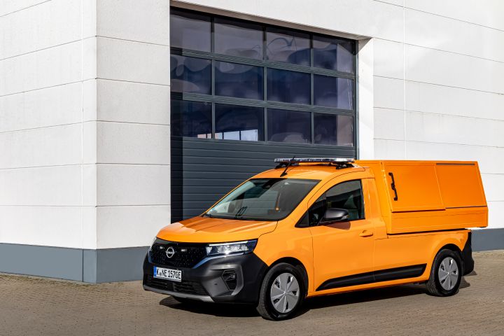 Vor einer hellen Wand mit großem Tor steht ein leuchtend oranger Nissan Townstar EV mit Sammelbehälter-Aufbau