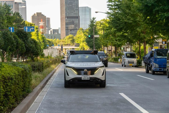 Ein weißer Nissan Ariya fährt in der Mitte der Straße und wird von vorne fotografiert. Im Hintergrund sind Hochhäuser und Bäume zu sehen, zusammen mit anderen Fahrzeugen, die auf beiden Seiten der Straße fahren.