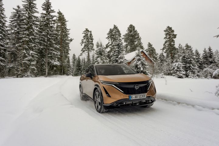 Elektroauto Nissan Ariya e‑4ORCE in Akatsuki Copper mit Dach und Außenspiegel in Pearl Black fährt auf schneebedeckter Straße mit verschneitem Wald und Holzhaus im Hintergrund, 3/4 Front
