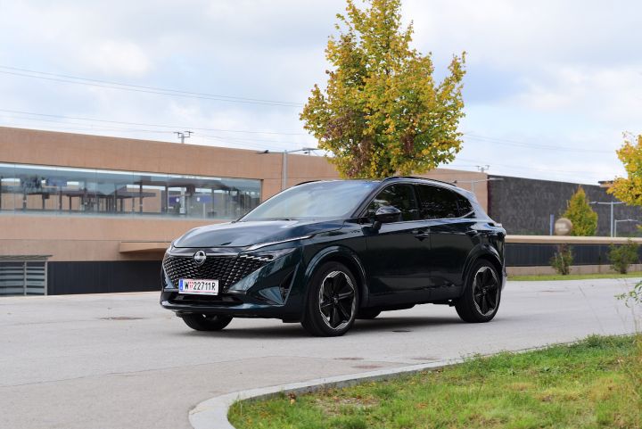 3/4 Frontansicht eines dunkelgrünen Nissan Qashqai e‑Power, der vor einem modernen Gebäude mit braun-schwarzer Fassade steht