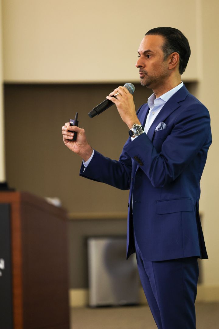 A man in a blue suit speaking into a microphone with a small device in his other hand.