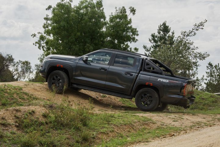 2026 Nissan Navara PRO-4X finished in Boulder Grey side‑on while climbing an earthen berm, showing extended suspension, sports bar, black alloys and orange arch tabs.
