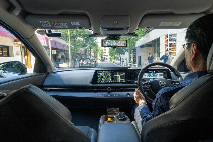 Blick aus dem Inneren eines Nissan Ariya. Eine Person sitzt auf dem Fahrersitz und blickt nach vorne, wobei das Armaturenbrett und das Navigationssystem zu sehen sind. Im Hintergrund sind Straßen mit Gebäuden und Fußgängern zu sehen.