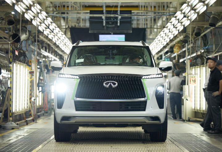A white all-new 2025 QX80 SUV progresses towards the end of the production line down as three workers stand to the left.