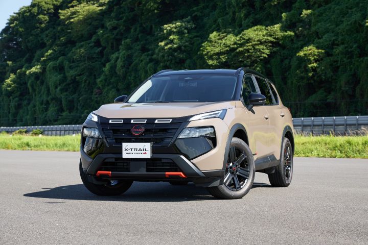 A beige X‑Trail is parked on a paved road. The vehicle is facing slightly to the left, and the Nissan logo is visible on the front grille. The license plate reads 'XTRAIL', and lush green trees fill the background.
