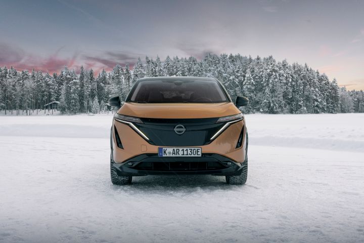Elektroauto Nissan Ariya e‑4ORCE in Akatsuki Copper mit Dach und Außenspiegel in Pearl Black auf Eissee mit schneebedeckten Bäumen im Hintergrund, Front