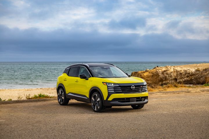 Front right 3/4 view of a neon yellow 2025 Nissan Kicks parked on a gravel lot overlooking the ocean with thick clouds above.