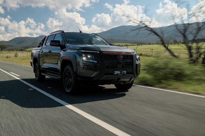 2026 Nissan Navara PRO-4X finished in Boulder Grey shown in a side-leading tracking frame with blurred foreground twigs, black wheels and mountain plain.