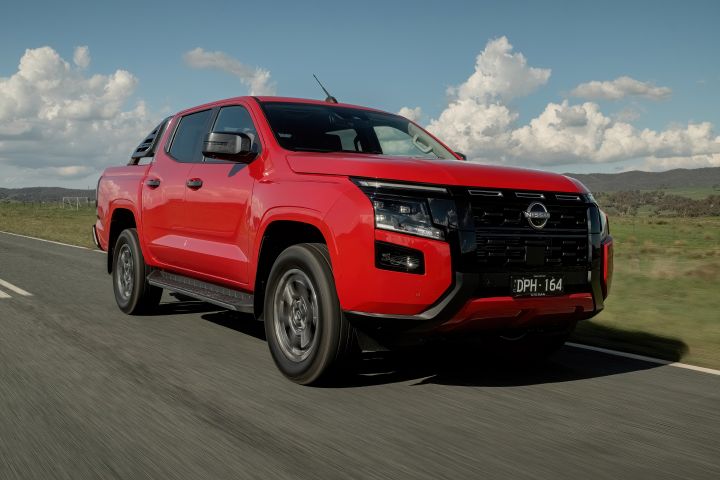 Front three‑quarter view of a 2026 Nissan Navara ST‑X dual cab in Outback Red, driving on a sealed road with no incline, surrounded by roadside vegetation and mountains.