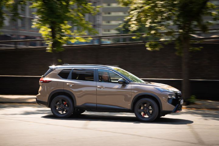 Champagne-coloured Nissan X‑Trail driving through an urban area, shown in side profile with trees and city buildings in the background.