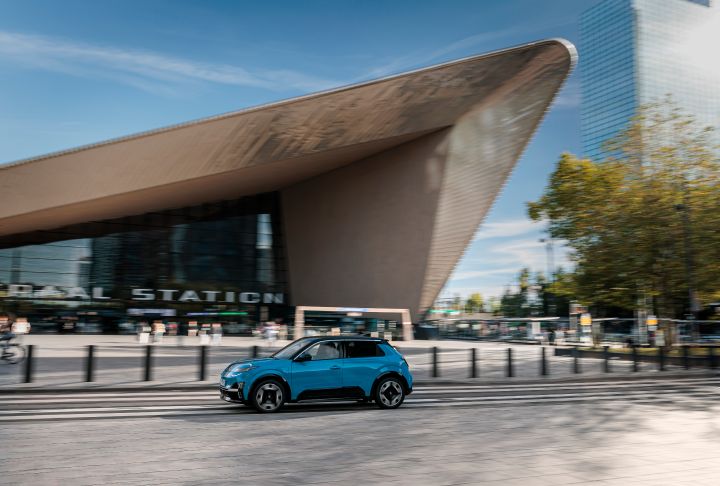 Ein blauer Nissan Micra fährt auf einer Straße vor dem Rotterdamer Hauptbahnhof mit auffälliger, spitz zulaufender Dachlinie vorbei